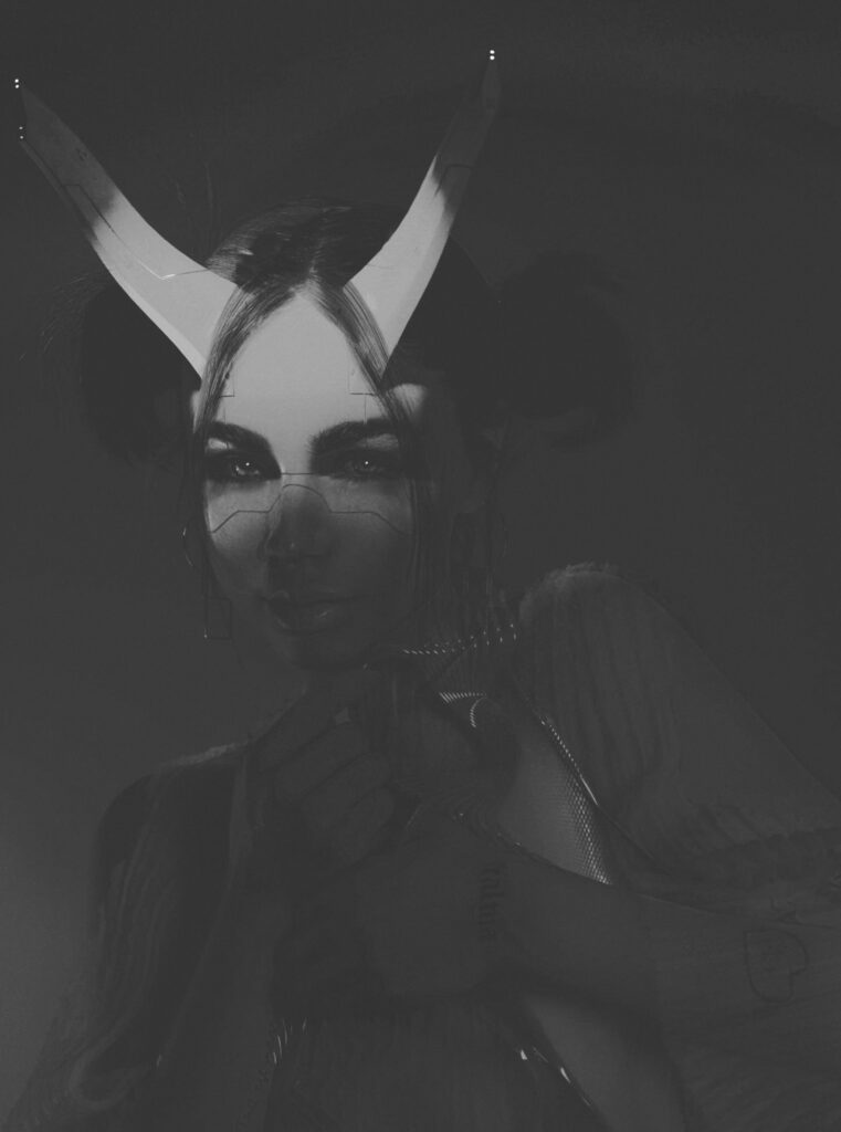 Darian Rojas with horns and intricate makeup stands against a gradient backdrop wearing a translucent outfit with layered wavy designs.